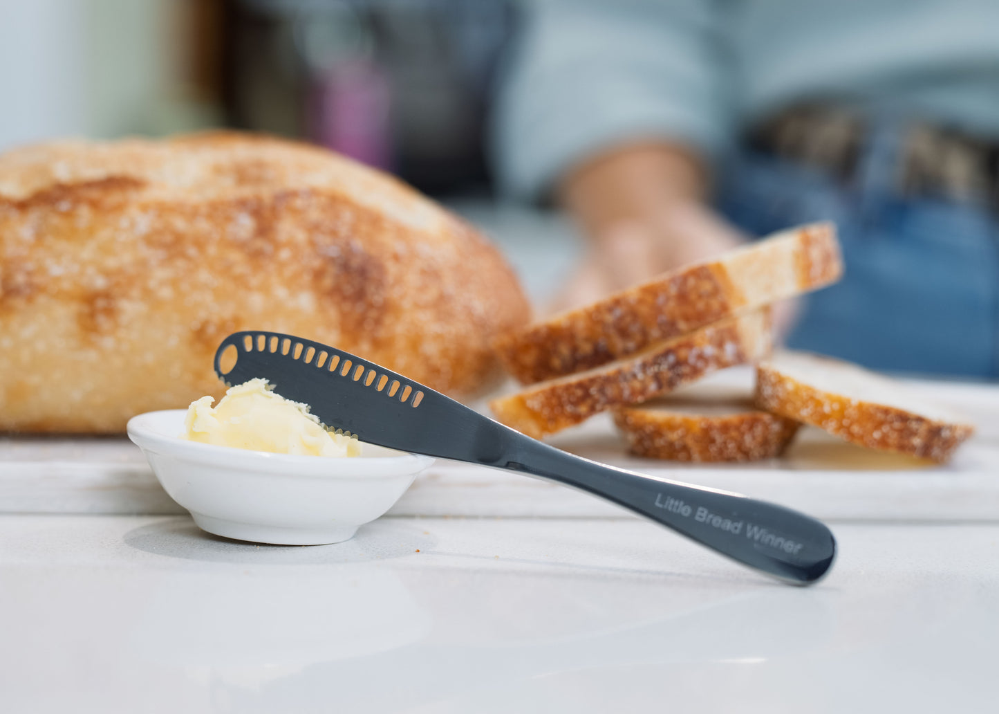 A stainless steel butter spreader knife with the words 'Little Bread Winner' printed on the handle, resting on a white dish with butter, with a loaf of bread and sliced bread in a blurred kitchen background.