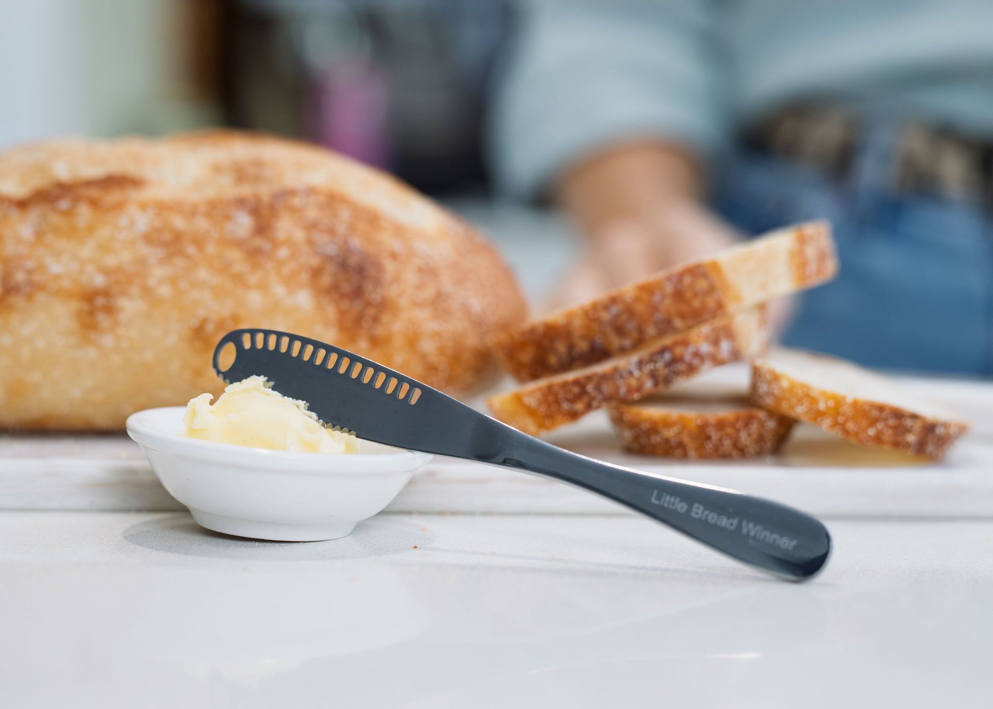 A stainless steel butter spreader knife with the words 'Little Bread Winner' printed on the handle, resting on a white dish with butter, with a loaf of bread and sliced bread in a blurred kitchen background.