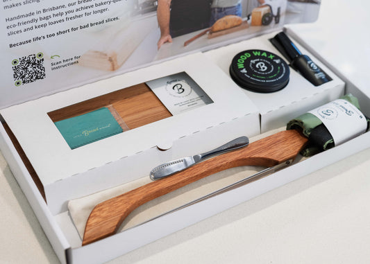 An open white gift box containing a wooden bread board  and timber bread saw, a rolled green waxing cloth, a round black tin of wood wax , a butter knife, and rolled bread bag