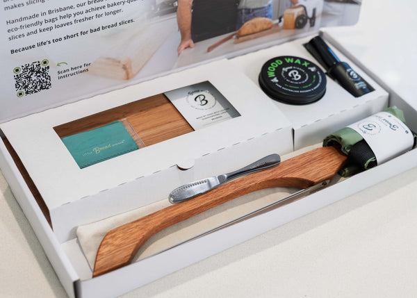 An open white gift box containing a wooden bread board  and timber bread saw, a rolled green waxing cloth, a round black tin of wood wax , a butter knife, and rolled bread bag