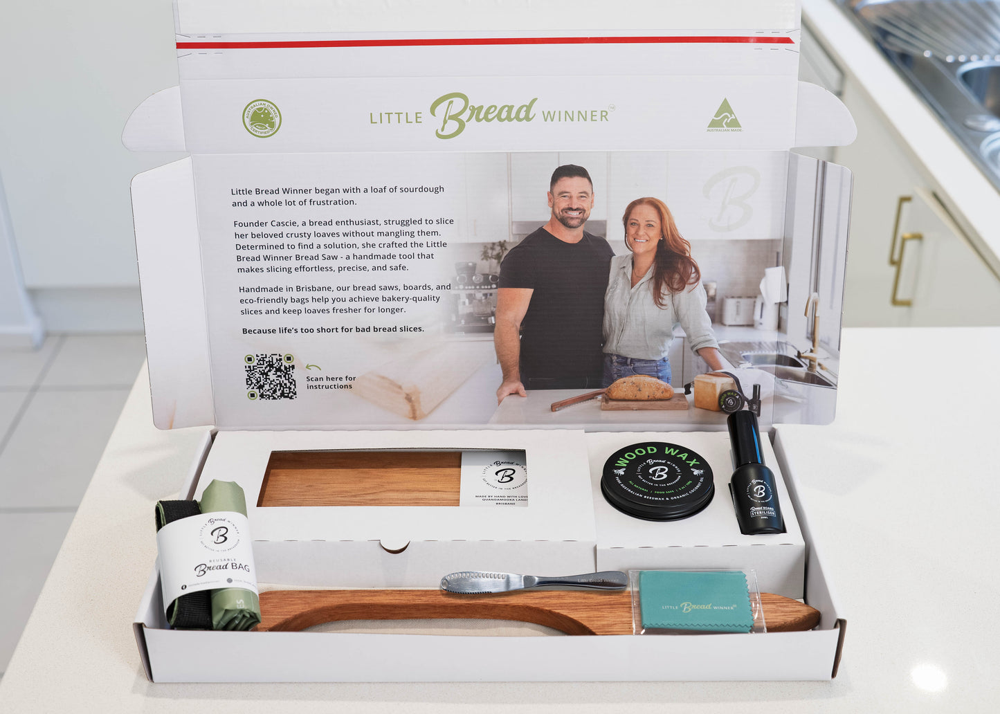 An open white gift box containing a wooden bread board  and timber bread saw, a rolled green waxing cloth, a round black  tin of wood wax , a butter knife, and rolled bread bag