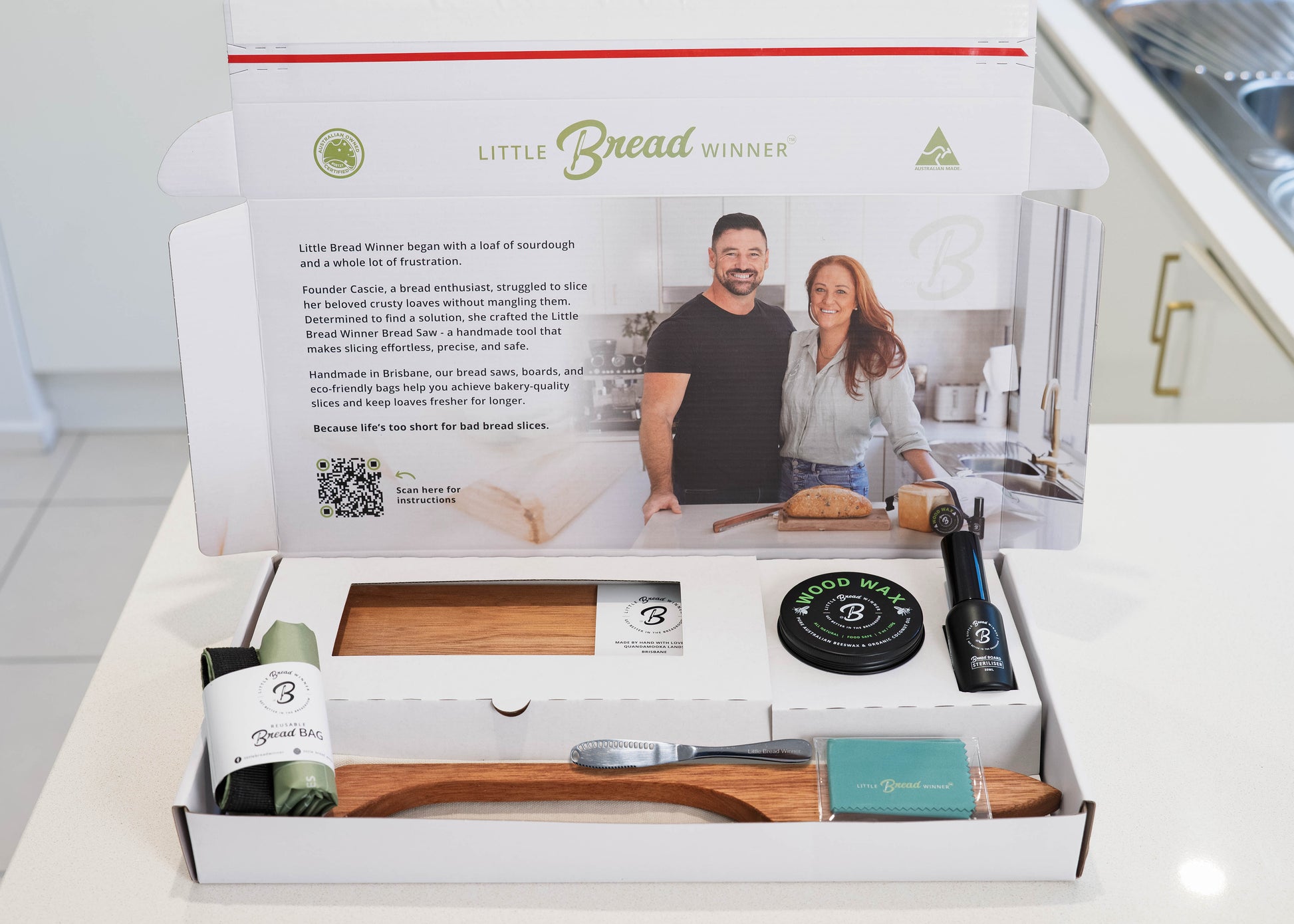 An open white gift box containing a wooden bread board  and timber bread saw, a rolled green waxing cloth, a round black  tin of wood wax , a butter knife, and rolled bread bag