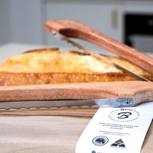 Wooden bread saw and cutting board with a loaf of bread in the background on a kitchen counter.