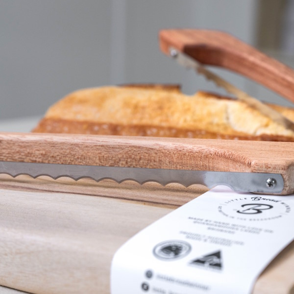 Close focus on wooden bread saw and timber cutting board with loaf of bread in the background on a kitchen counter.